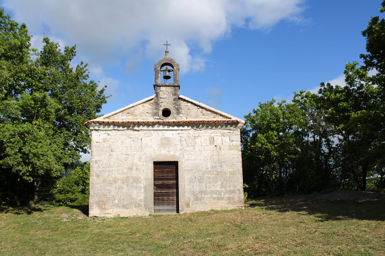 A rustic stone chapel stands alone in a peaceful forest clearing on a sunny day.